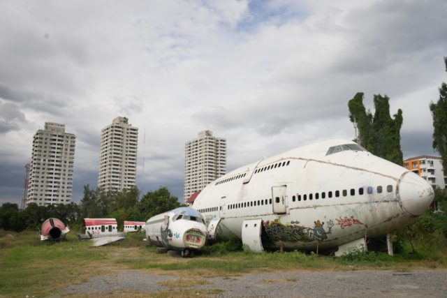 Two abandoned and damaged planes sit side by side on the ground, buildings in the distance.