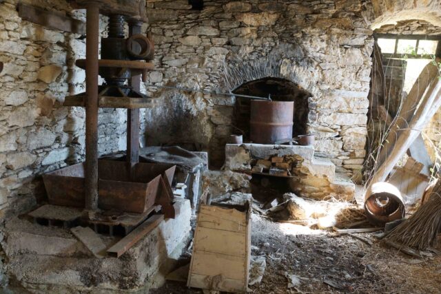 Inside of a stone building, a hearth and shelving stand whiles debris litters the ground.