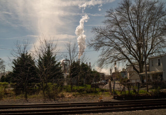 Smoke coming out of a crude oil plant surrounded by houses and trees.