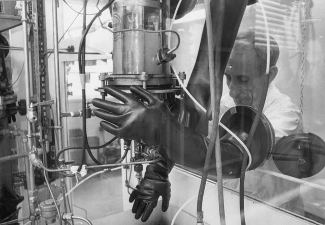 Scientist wearing gloves behind a glass shield.