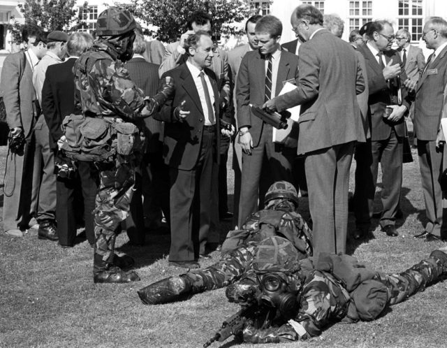 Group of men in suits stand around talking while two men in camouflage uniforms lie on the ground.