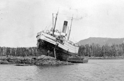 Large steamship wrecked on a rocky island with water in the foreground and trees in the background.