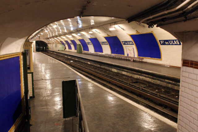 View looking down into Porte des Lilas station with white walls, station signs, and frames painted blue along the walls.