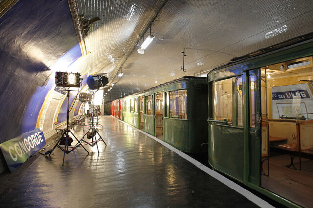 View of an old green Metro train in a station with film lights on the platform.
