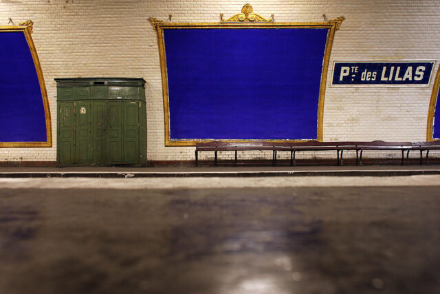 Métro station wall with a green cabinet, benches, large picture frames painted blue, and a sign that reads "Pte. des Lilas."