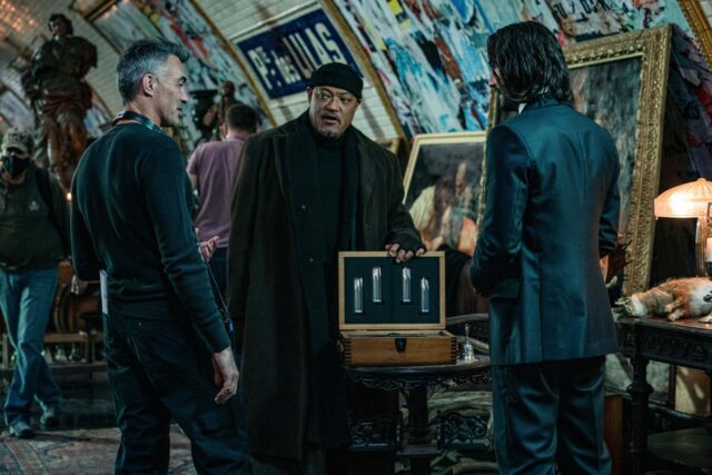 Keanu Reeves, Laurence Fishburne, and Chad Stahelski standing with crew in the Porte des Lilas Métro station surrounded by paintings. 