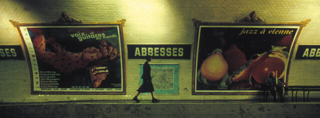 Audrey Tautou in chunky boots and all black walks across a Métro platform with two large painting behind her and a sign that reads "Abbesses."