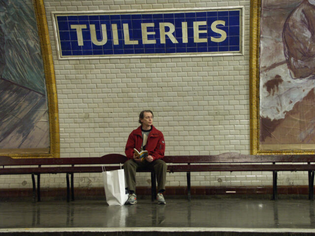 Steve Buscemi sitting at a bench in a Métro station wearing a red jacket with a sign that reads "Tuileries," behind him.