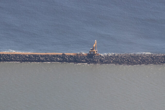 Excavator on a rock jetty in the water.