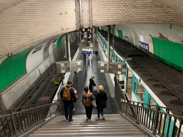 People walking down a set of stairs into the Porte des Lilas Métro station with green paintings on either side and a train with open doors on the right.