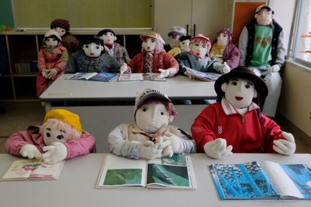 Dolls that look like children sitting at two school desks. 