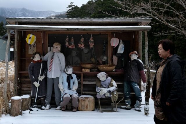 Group of dolls standing and sitting in a wooden shelter with Tsukimi Ayano walking in front.