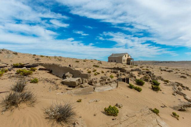Abandoned building in the distance with shrubs and rocks in the sand in the foreground. 
