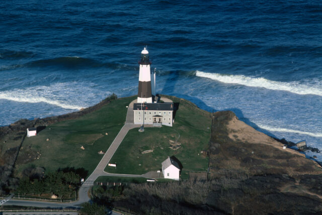 An aerial view of a lighthouse on the edge of land, water on the other side.