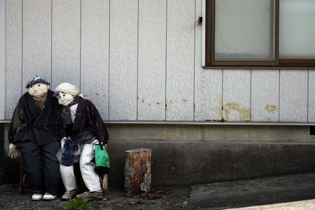 Dolls made to look like an elderly couple lean against each other in front of a wall. 
