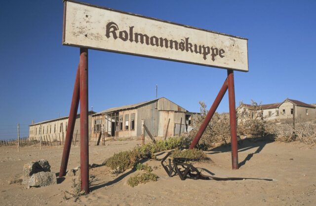 Sign reading "Kolmannskuppe" standing in front of a numbers of abandoned buildings. 