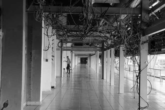 A hallway with exposed wires in the ceiling, a man walking