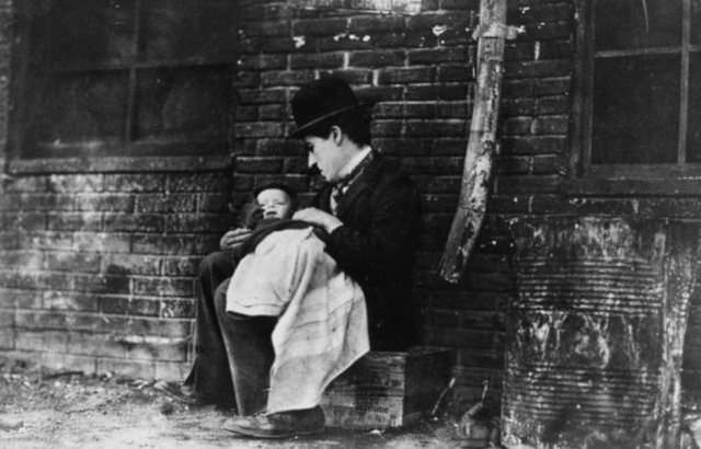 Charlie Chaplin sits on a box holding a baby in a scene from The Kid.
