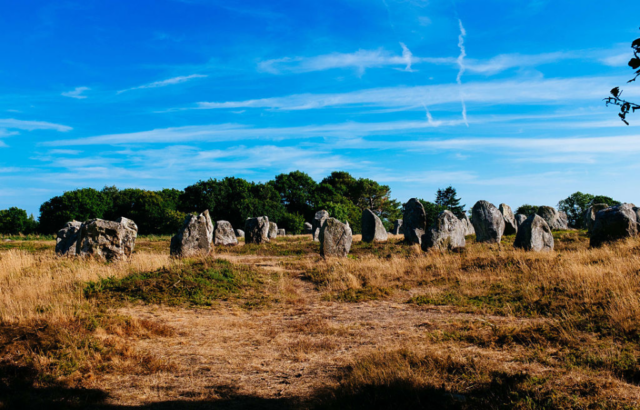 View looking out at standing stones in France across a field of dried grass with a blue sky.
