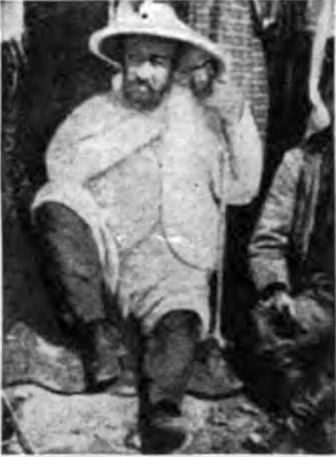 An old photo of an archaeologist sitting down, holding his hat, one knee bent