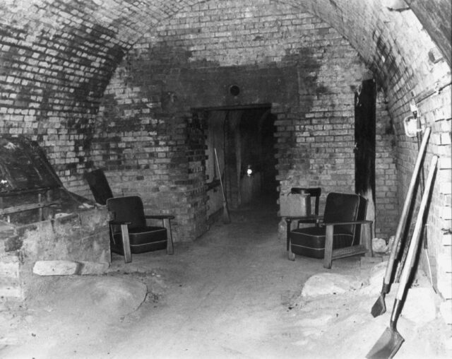 Two chairs and some wooden crates inside an underground brick bunker.