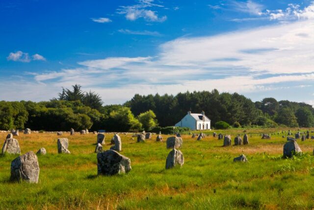 View across a green field full of stone monoliths looking towards a small white house, with a blue sky in the background. 