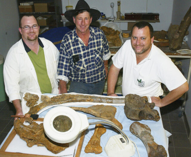 Three men pose for a photo behind a table of dinosaur fossils.