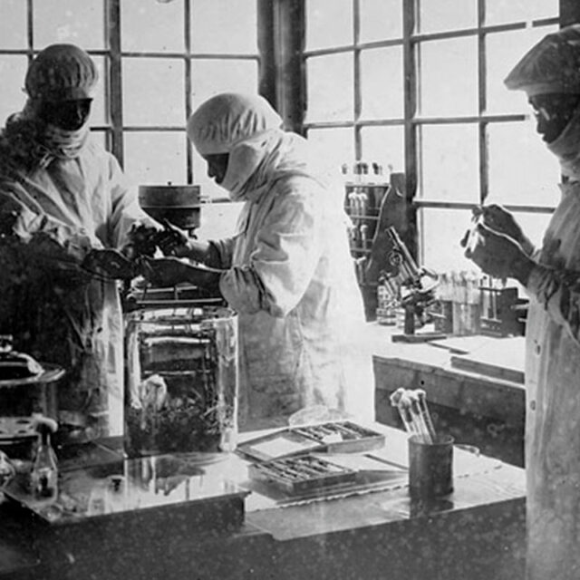 Three members of Unit 731 standing around laboratory equipment while dressed in protective gear