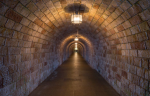 Look down a long brick tunnel lit by overhead lights, with three figured in the distance.