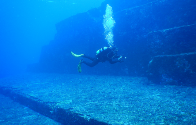 A scuba diver swims past a stepped stone structure underwater.