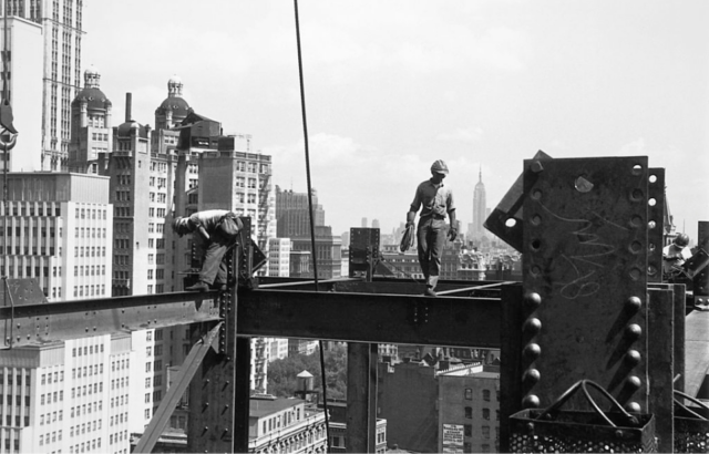 Two construction workers walk on metal beams high in the air.