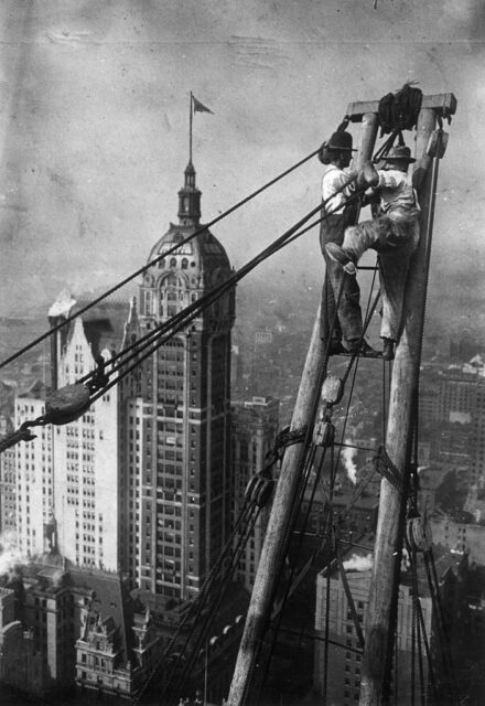 Two workers stand on a metal rung beside a series of cables, high above a city.