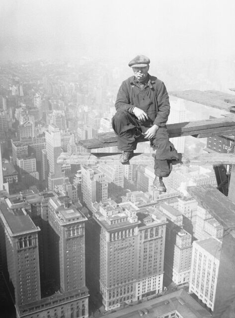 Man sitting on a piece of wood high above a cityscape. 