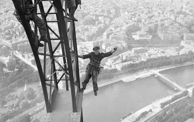 Man standing on a metal platform of the Eiffel Tower leaning out to the side. 