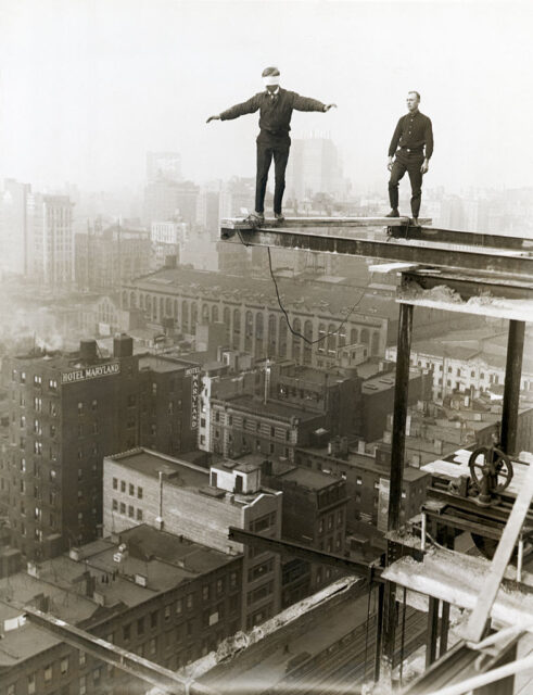 Blindfolded man walks across a wooden beam high above a cityscape while his friend watches on.