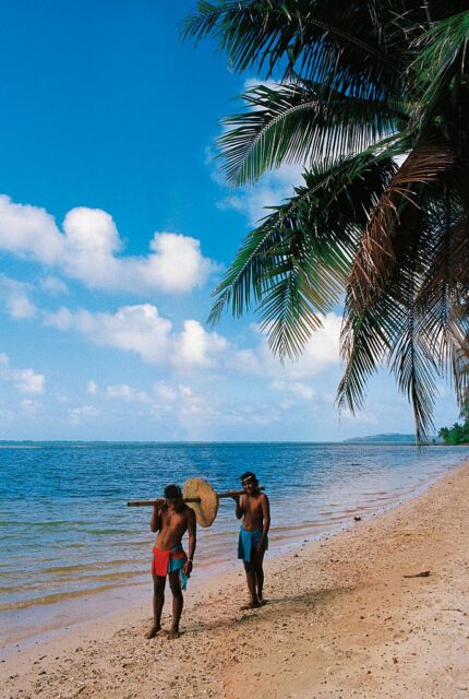 Two shirtless men carry a small stone coin on a stick over their shoulders.