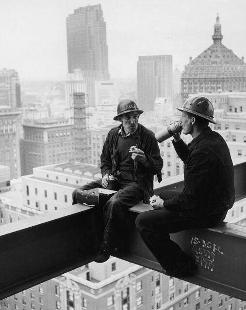 Two construction workers with hard hats on sit on scaffolding while they eat their lunches.