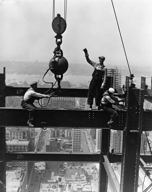 Two men sit on a metal beam high above a city, while another stands to direct a crane into position.