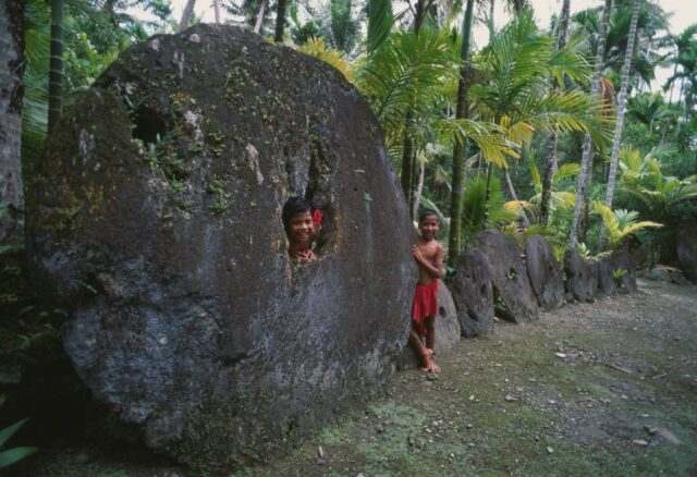 Two children in loin cloths stand with a tall circular stone with a small circle cut in the center.