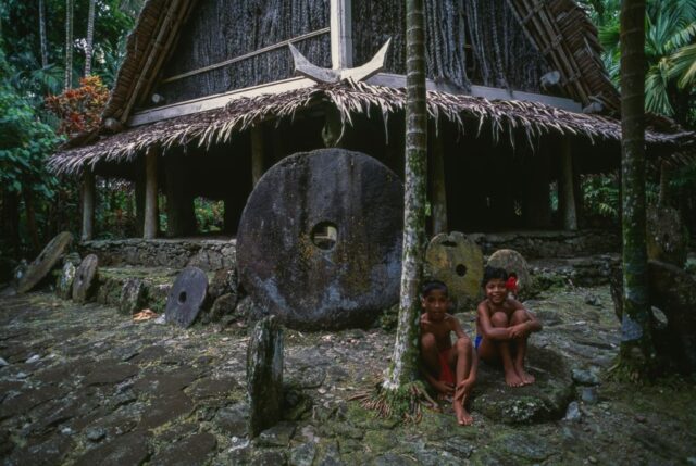 Two children sitting beside a large stone circle with a small circle cut in the middle, and a wood hut in the background.