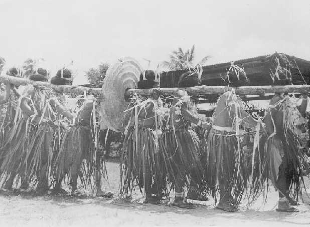 Yapese villagers in grass skirts hold a large circular stone held up by a branch through it resting on their shoulders.