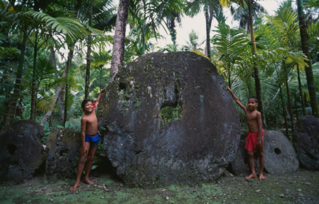 Two children in loin cloths stand beside a large circular stone with a small hole in the middle.