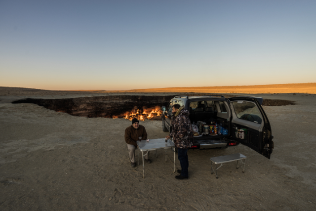 Two people with a table are hanging out behind a vehicle with open hatch, flanked by Turkmenistan's Gates of Hell.