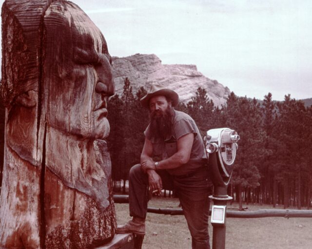 Korczak Ziolkowski leaning on his knee in front of a carving of Crazy Horse. The Crazy Horse Memorial mountain in the background.