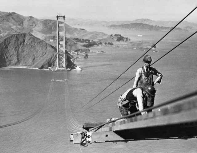 Workers building a catwalk on the Golden Gate Bridge, view of the unfinished bridge behind them.