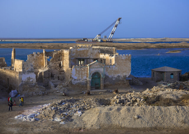 People walking toward a semi-demolished building, a lake in the background with construction equipment.