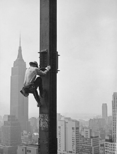 A construction worker climbing a steel pole in the sky.