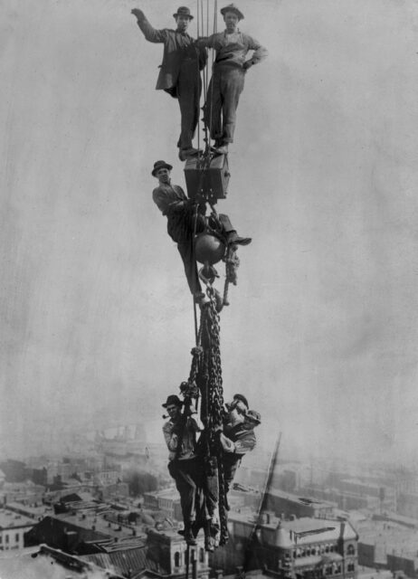 Several construction workers hanging off a crane's chain.