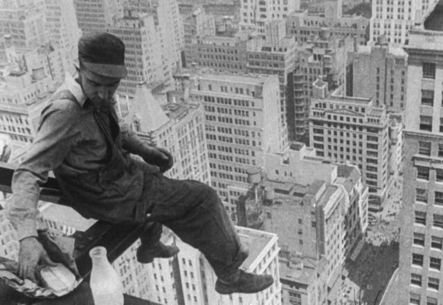 A construction worker sitting on the edge of a steel girder high in the sky.