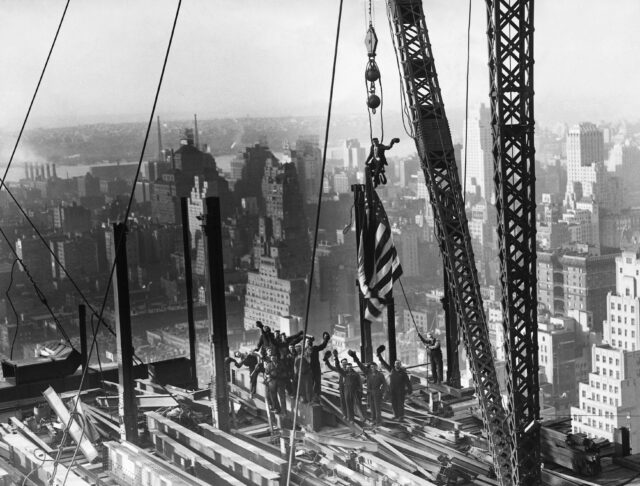 A group of construction workers salute the camera with their hats on a skyscraper construction site.